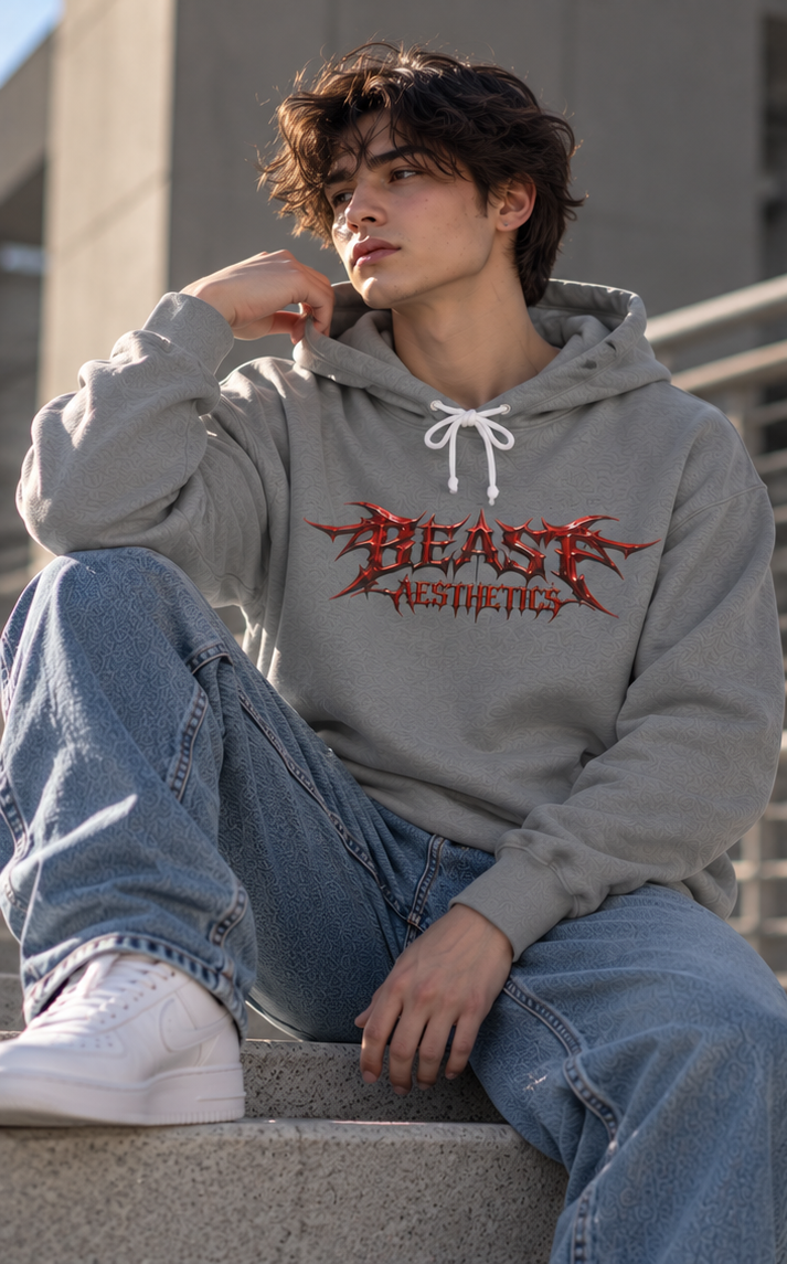 A young man wearing a light gray hoodie with a red and black 'BEAST AESTHETICS' graphic, paired with blue jeans and white sneakers, sitting on concrete steps.