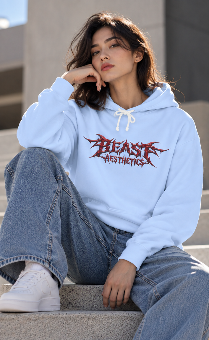 A woman sitting on concrete steps wearing a light blue hoodie with a red and black 'BEAST AESTHETICS' graphic on the chest, paired with wide-leg blue jeans and white sneakers.
