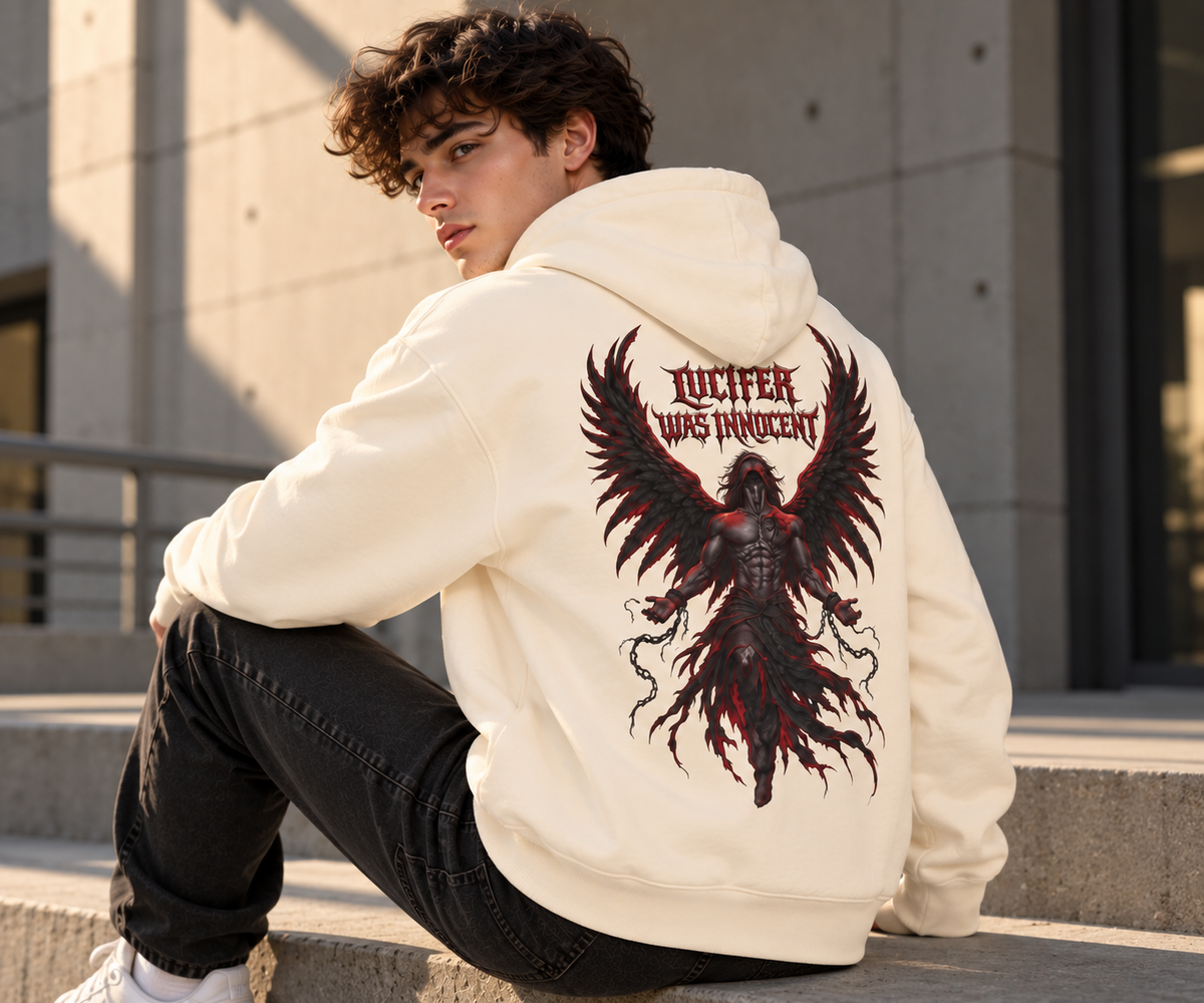 A young man sitting on outdoor concrete steps, wearing a cream-colored hoodie with a large graphic print on the back. The graphic features a dark angel with red wings and the text 'LUCIFER WAS INNOCENT'.