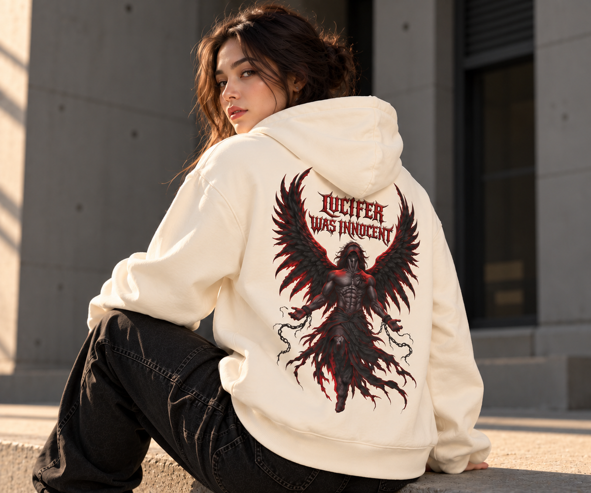 A woman wearing a cream-colored hoodie with a large black and red graphic of a winged demon and the text 'LUCIFER WAS INNOCENT' on the back, sitting outdoors against a concrete wall.