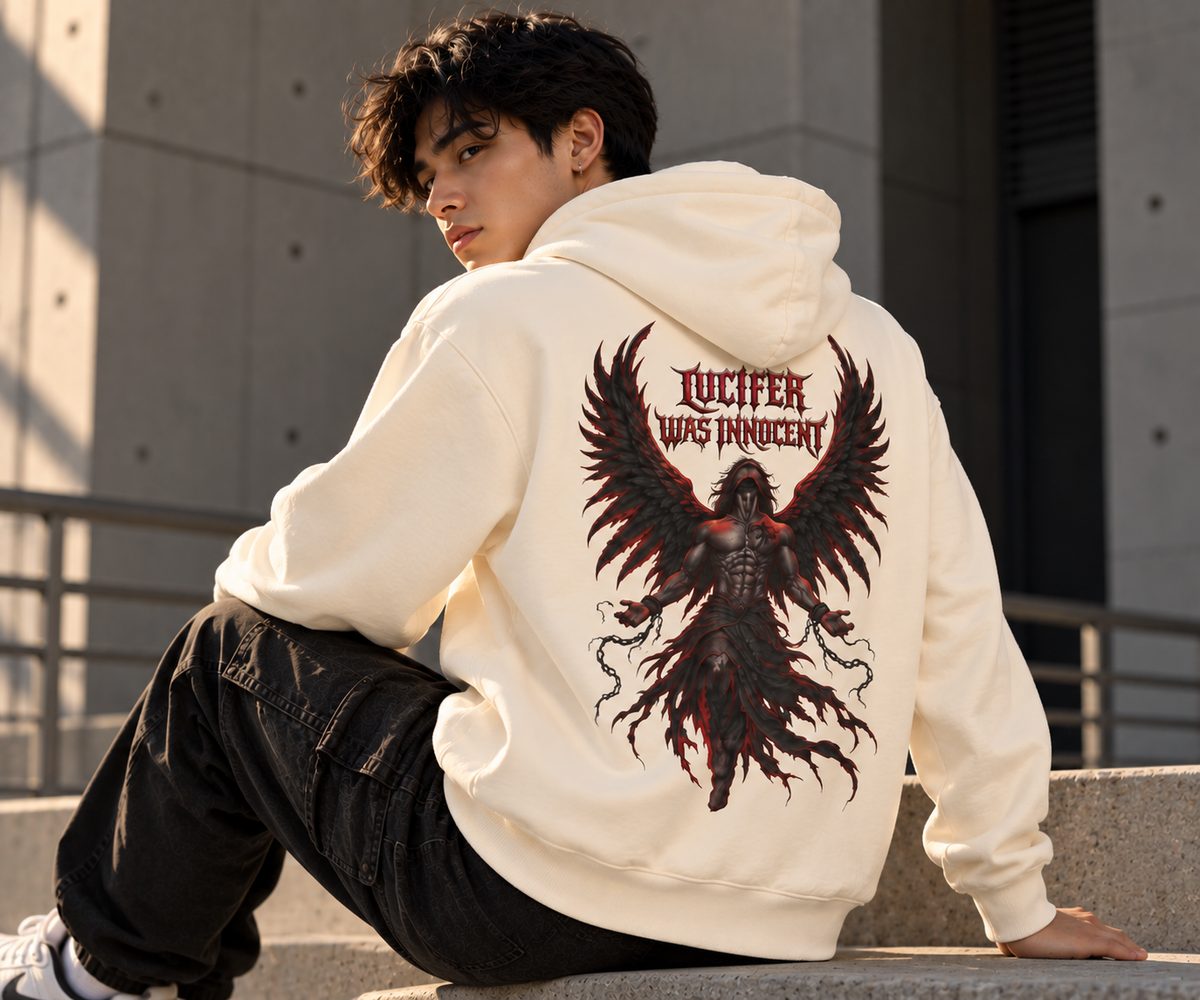 A man sitting on concrete steps outdoors, wearing a cream-colored hoodie with a large back graphic of a dark angel with wings and the text 'LUCIFER WAS INNOCENT'.