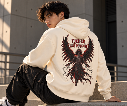 A man sitting on concrete steps outdoors, wearing a cream-colored hoodie with a large back graphic of a dark angel with wings and the text 'LUCIFER WAS INNOCENT'.